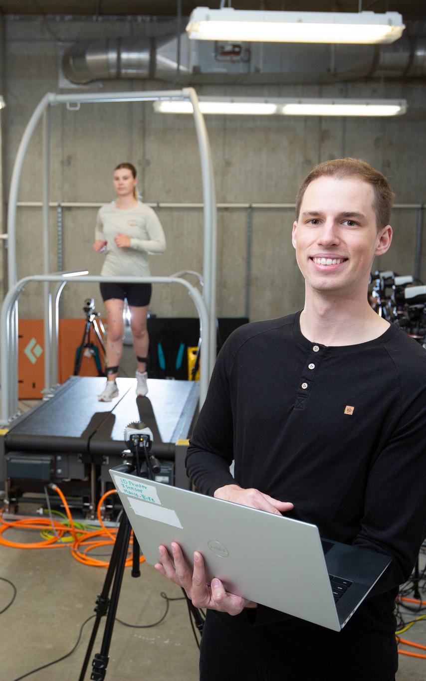 Smiling young man with laptop with young woman behind him running a treadmill